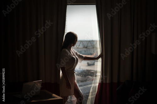 Young pretty woman standing by a window looking outside expecting someone. people and hope concept - close up of happy woman opening window curtains Caucasian model standing at window with city