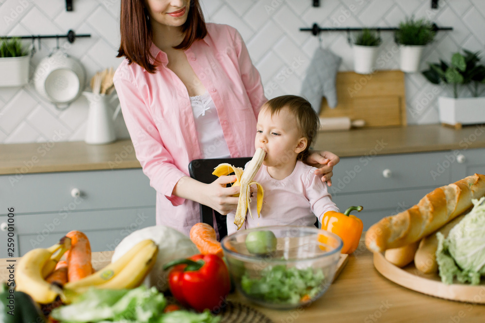Happy loving family, mother and daugher in the kitchen. Beautiful young mother feeding her cute baby girl with a banana at kitchen. Little baby girl eats banana sitting at the table