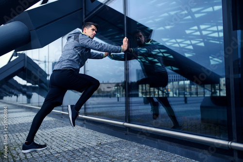 Sports man doing his stretching workut in scenery of modern city architecture. Stylish sportswear.
