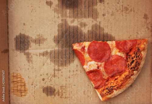 Pizza box with single slice of old pepperoni pizza in delivery box left after the party. Cold pizza leftovers in greasy cardboard, top view from above