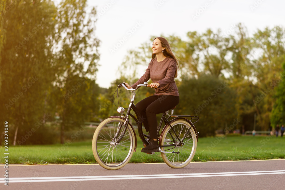 Young attractive woman riding through the park after work. Beautiful lady cycling during sunset. Bike as a trendy transport. Healthy outdoors activity on a warm summer day. Bicycle trend in the city.