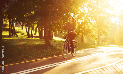 Young attractive woman riding through the park after work. Beautiful lady cycling during sunset. Bike as a trendy transport. Healthy outdoors activity on a warm summer day. Bicycle trend in the city. © Cressida studio