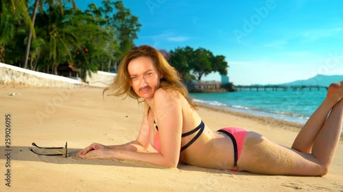 The concept of strange adventures of people. A beautiful woman in a pink bikinis on the beach, turned to the camera, and her mustache is visible on her face. photo
