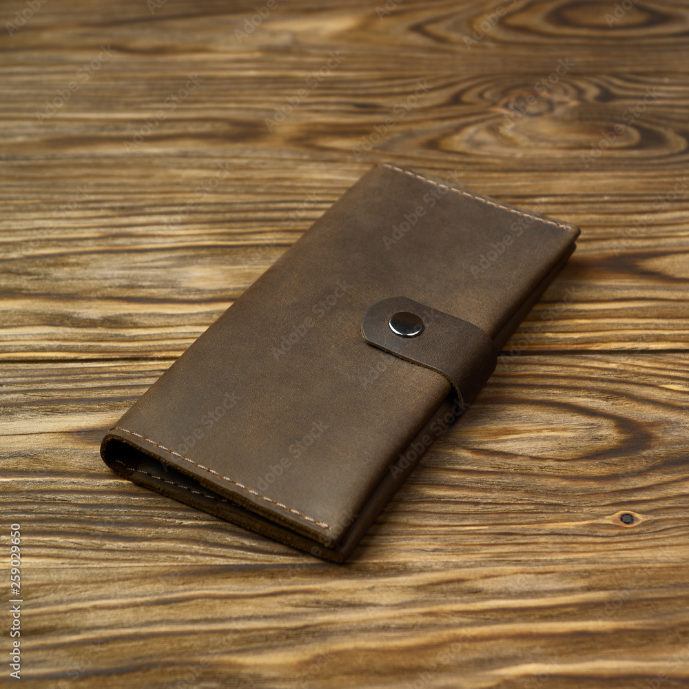 Brown color handmade leather wallet on textured wooden background. Wallet is unisex. Side view. Stock photo of luxury accessories. Brown color handmade leather wallet on textured wooden background. Wallet is unisex. Side view. Stock photo of luxury accessories.