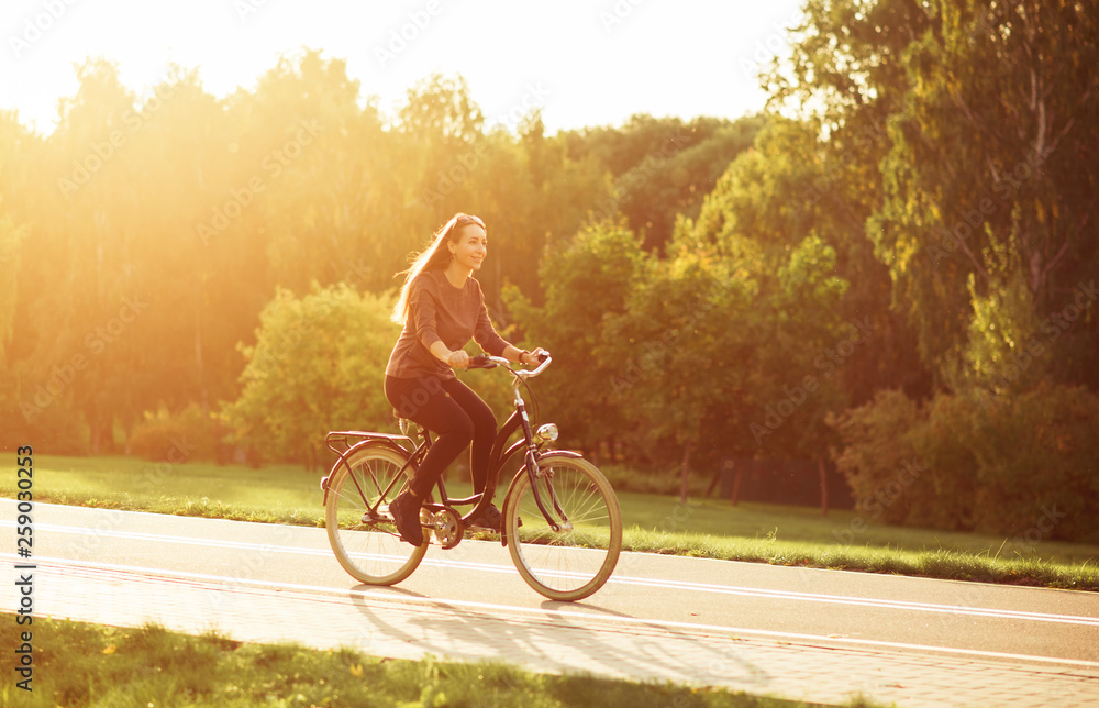 Obraz premium Young attractive woman riding through the park after work. Beautiful lady cycling during sunset. Bike as a trendy transport. Healthy outdoors activity on a warm summer day. Bicycle trend in the city.