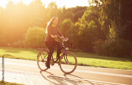 Fototapeta Naklejka Na Ścianę i Meble -  Young attractive woman riding through the park after work. Beautiful lady cycling during sunset. Bike as a trendy transport. Healthy outdoors activity on a warm summer day. Bicycle trend in the city.