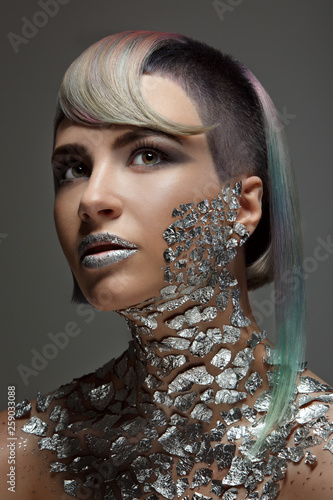 Beautiful girl with a short haircut and shaved temple and dyed hair. Metallic makeup and body painting, shiny pieces of metal spangles on the lips and chest. photo