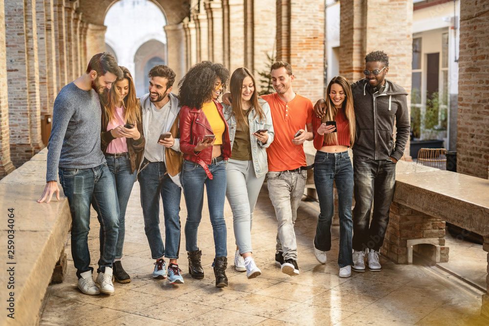 young adults converse and joke together using their smartphones walking under the arcades of a European city. Social media concept.