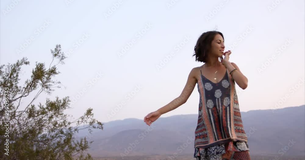 Stylish young hippy woman turns around to walk off frame in desert with mountain background Stylish young hippy woman turns around to walk off frame in desert with mountain background
