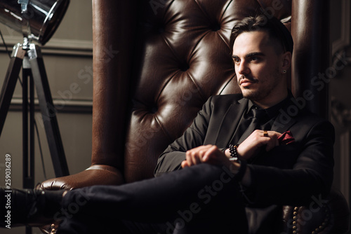 Young handsome man posing for a fashion shoot in a studio. Fashion as a lifestyle. Man wearing a beard. Model sitting on a sofa in tuxedo. Successful fashionable businessman. Business look advertising