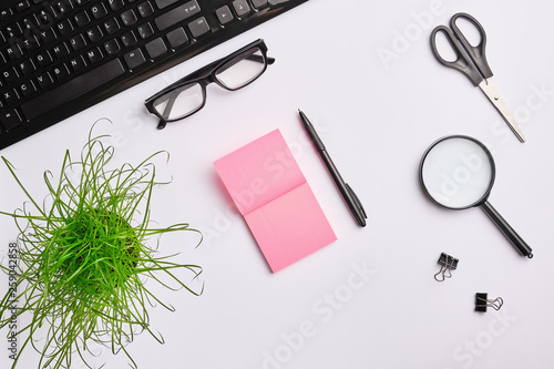 White table with keyboard, magnifying glass, stickers, scissors, office plant, clips and pen.