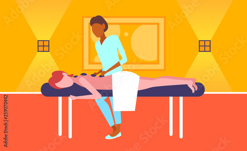 young girl enjoying traditional hot stone massage african american masseuse massaging patient woman hotel health spa concept horizontal full length