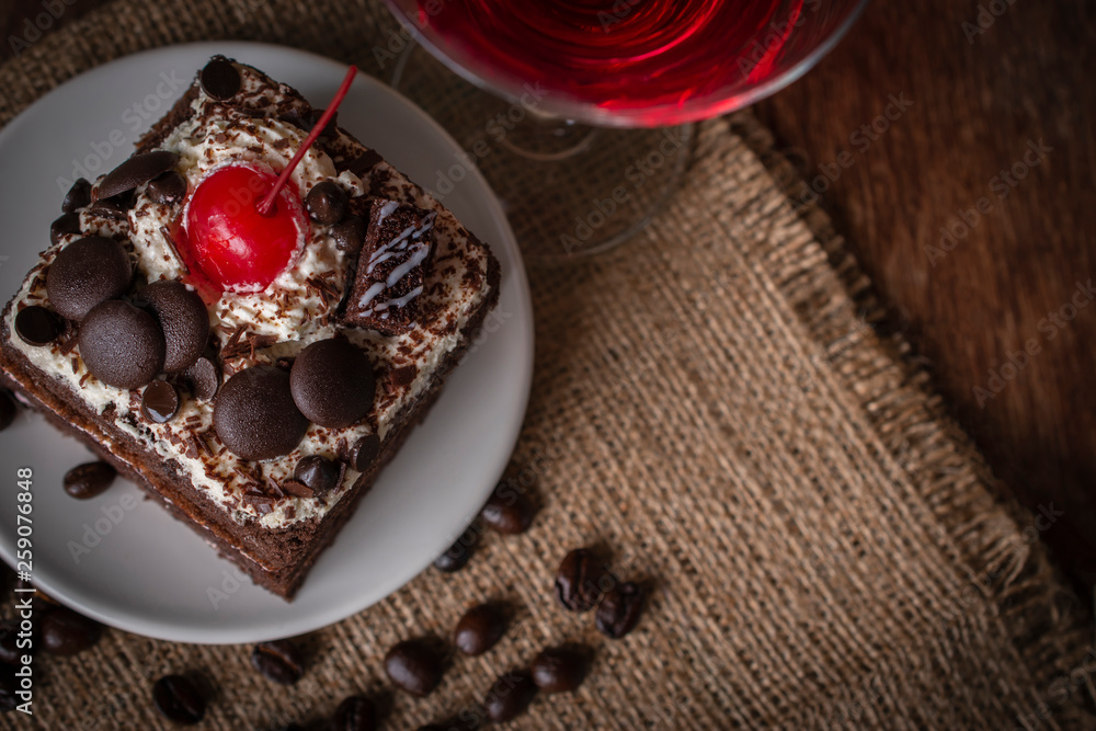 Black Forest Cake topping cherry putting on the white plate on the sack and wood background there are coffee bean and glass placed around. Black Forest Cake topping cherry putting on the white plate on the sack and wood background there are coffee bean and glass placed around.