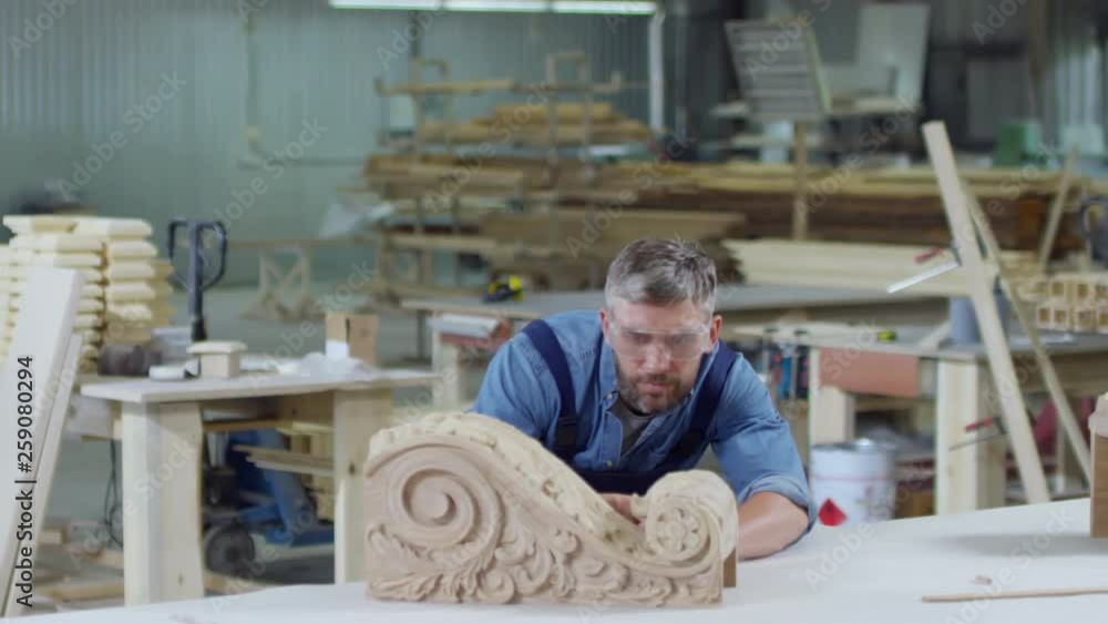 Panning of middle-aged Caucasian man in safety goggles and uniform sanding part of wooden bas-relief with sandpaper Panning of middle-aged Caucasian man in safety goggles and uniform sanding part of wooden bas-relief with sandpaper