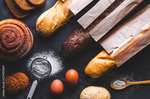 Baking ingredients for flour and rye bakery products. Fresh crisp bread, baguette packaged in paper bag and buns on a black background.