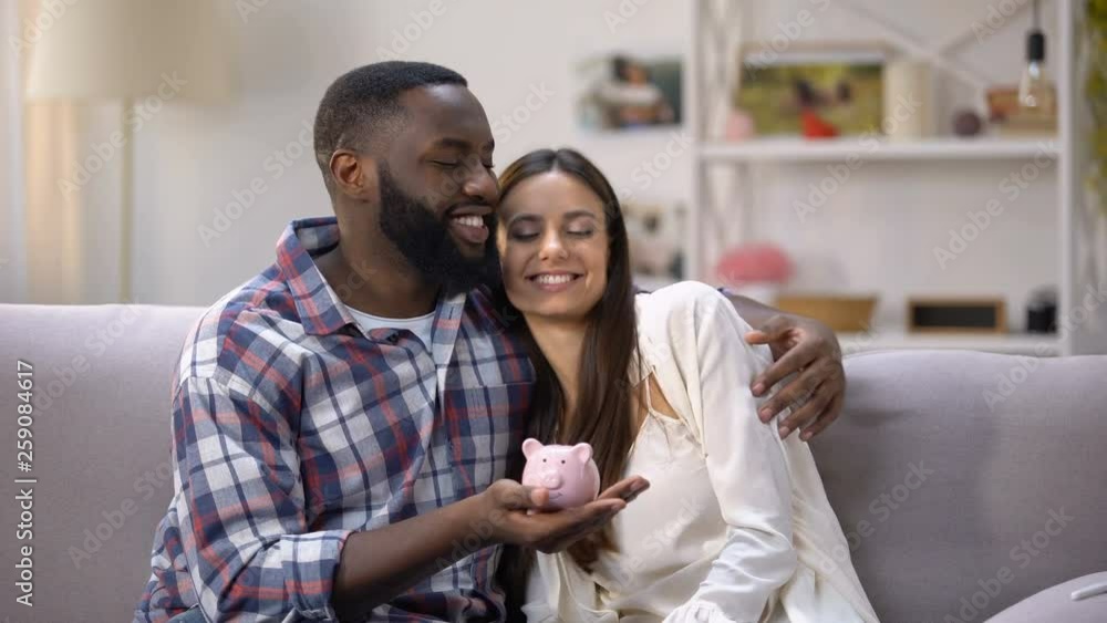 Mixed-race couple putting coins in piggy bank, social program for young families Mixed-race couple putting coins in piggy bank, social program for young families