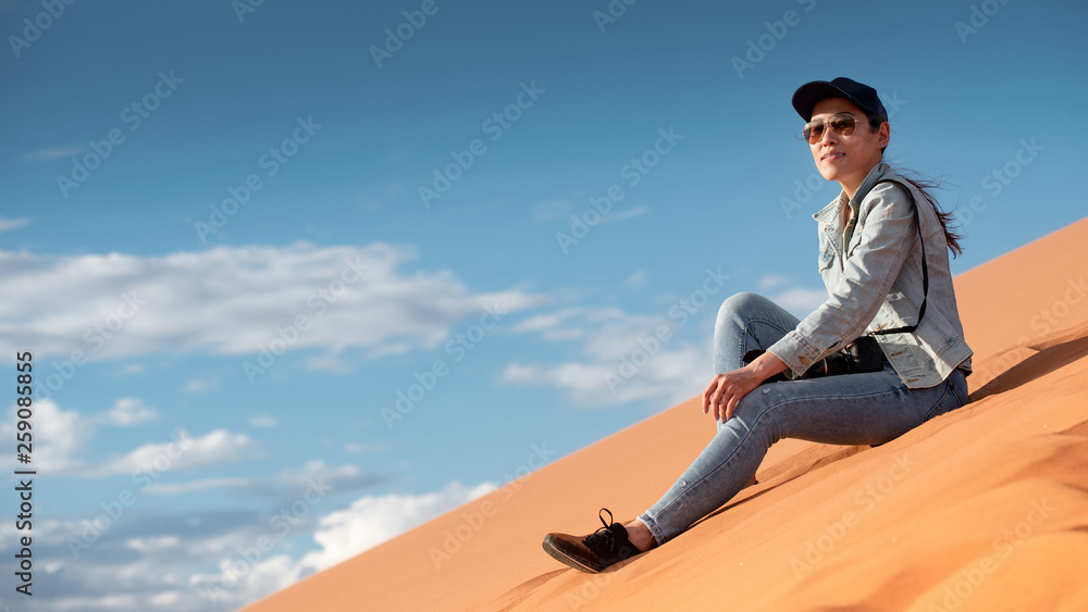 Young Asian woman traveler and photographer sitting on sand dune enjoy looking at the scenery in Namib desert of Namibia, Africa. Travel photography concept