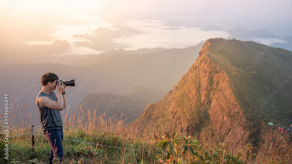 Young Asian male photographer and traveler taking photo of mountain landscape scenery during sunrise at Khao San Nok Wua in Kanchanaburi, Thailand. Travel photography concept