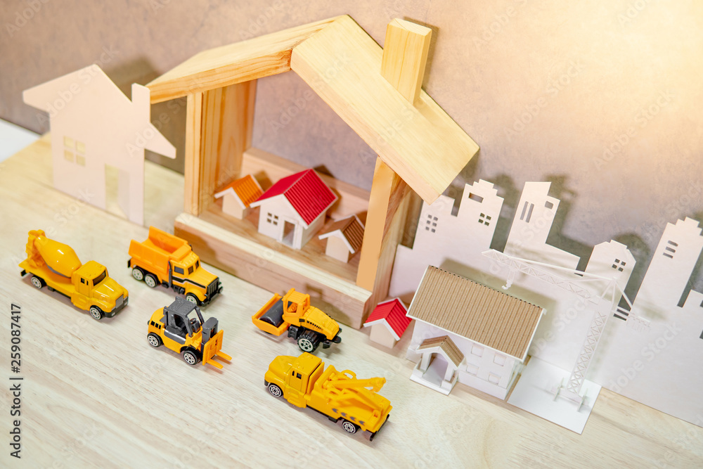 Obraz premium House models, paper crane and city background with miniature yellow trucks on wooden table. Architecture and construction industry for housing development business. Property or real estate concept