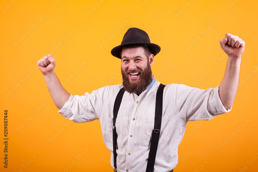 Attractive bearded man smiling and wearing a black hat rising his fists over yellow background Attractive bearded man smiling and wearing a black hat rising his fists over yellow background