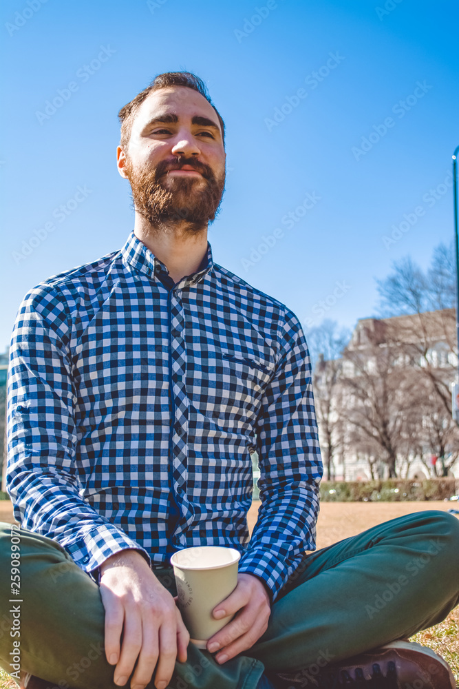 The young guy with beard and in shirt sits on grass having crossed legs in the spring park and has coffee from paper glass. The young guy with beard and in shirt sits on grass having crossed legs in the spring park and has coffee from paper glass.