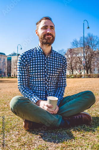 The young guy with beard and in shirt sits on grass having crossed legs in the spring park and drinks coffee from paper glass. Mood.