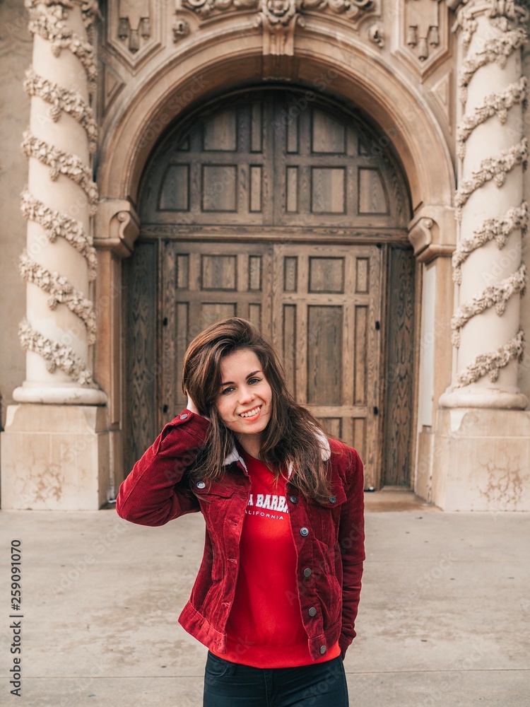 A brunette girl in a red jacket and with long hair stands at the door of a beautiful Museum building with stucco, arches and architecture in a Park in San Diego