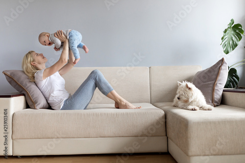 Young mother and child in her hands happy with white westie west highland white terrier dog on a white sofa with grey wallss miling and playng