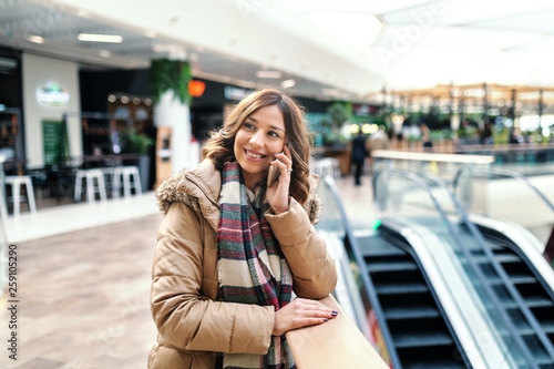 Smiling pretty brunette in coat and with scarf leaning on the fence and talking on smart phone while standing at shopping mall. © dusanpetkovic1