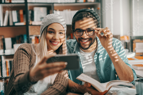Two multicultural hipster students taking selfie in library while sitting at desk. Man holding book and woman taking photo.