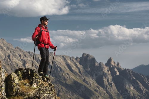 Porter of a bearded traveler hipster in sunglasses and a cap with a backpack and trekking poles stands on a high cliff near the cliff on the background of epic mountains of rocks and clouds