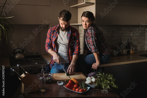 Beautiful young couple in kitchen at home while cooking healthy food. Man is cuts salad. Woman sits on table and looks in frame. Scene from family life.