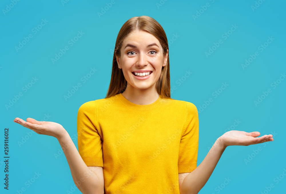 Smiling young girl looks at camera and spreads hands. Photo of attractive girl shows open palms with something in yellow sweater on blue background Smiling young girl looks at camera and spreads hands. Photo of attractive girl shows open palms with something in yellow sweater on blue background