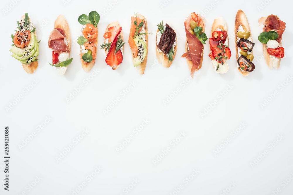 Fototapeta top view of traditional italian bruschetta with prosciutto, salmon, fruits, vegetables and herbs on white with copy space