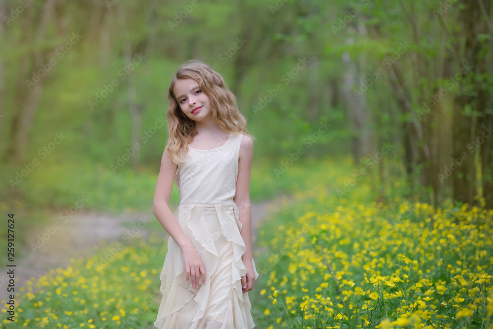 A cute young girl is in the forest on the edge of the forest with yellow flowers. She has blond curly hair, she's wearing a beautiful dress. A cute young girl is in the forest on the edge of the forest with yellow flowers. She has blond curly hair, she's wearing a beautiful dress.