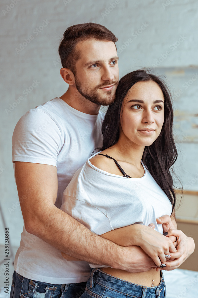 handsome man hugging beautiful and brunette woman in t-shirt and looking away