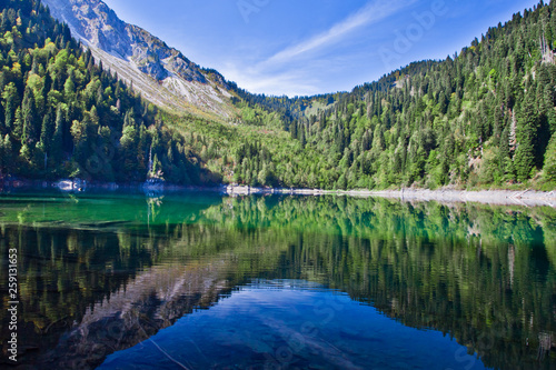 Fototapeta Naklejka Na Ścianę i Meble -  Symmetrical reflections. Turquoise water of a mountain lake surrounded by green wooded hills under a blue sky. Lake Ritsa Tourism in the Caucasus in Abkhazia.