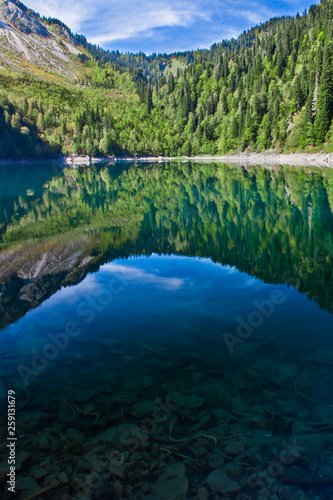 Symmetrical reflections. Turquoise water of a mountain lake surrounded by green wooded hills under a blue sky. Lake Ritsa Tourism in the Caucasus in Abkhazia.