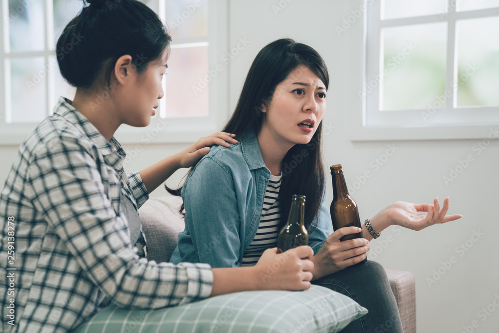 Fototapeta premium asian woman complaining telling to friend about her problems sitting on couch sofa at home. two girls holding bottle of beers drinking. roommate concentrated listening to lady and comforting her.