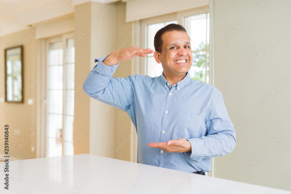 Middle age man sitting at home gesturing with hands showing big and large size sign, measure symbol. Smiling looking at the camera. Measuring concept. Middle age man sitting at home gesturing with hands showing big and large size sign, measure symbol. Smiling looking at the camera. Measuring concept.