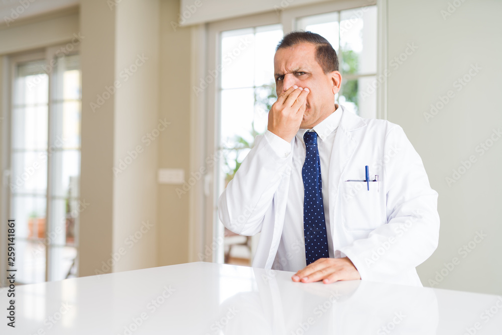 Middle age doctor man wearing medical coat at the clinic smelling something stinky and disgusting, intolerable smell, holding breath with fingers on nose. Bad smells concept.