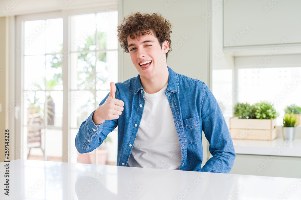 Young handsome man wearing casual denim jacket at home doing happy thumbs up gesture with hand. Approving expression looking at the camera showing success.