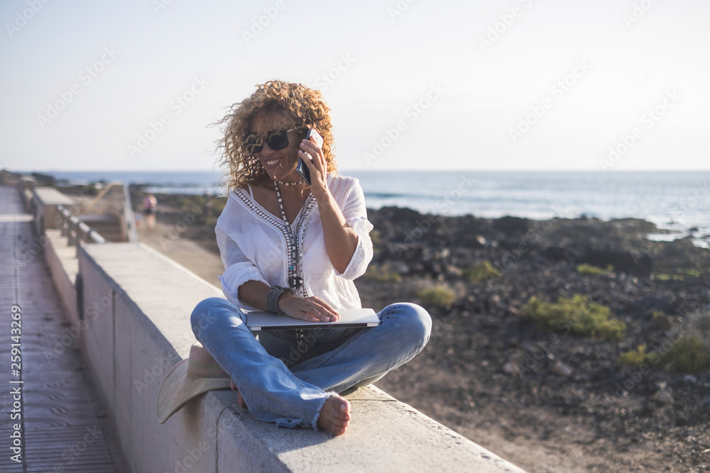 Young beautiful caucasian woman call at the phone speaking at the beach - laptop on his legs for modern trtaveler technology concept - summer vacation with internet connection outside