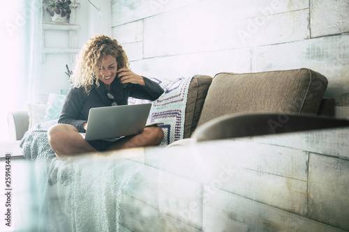 Curly woman with gray sweater sitting on brown sofa and blue blankets relaxes looking at the computer on an autumn day. Technology concept for independent people enjoying at home