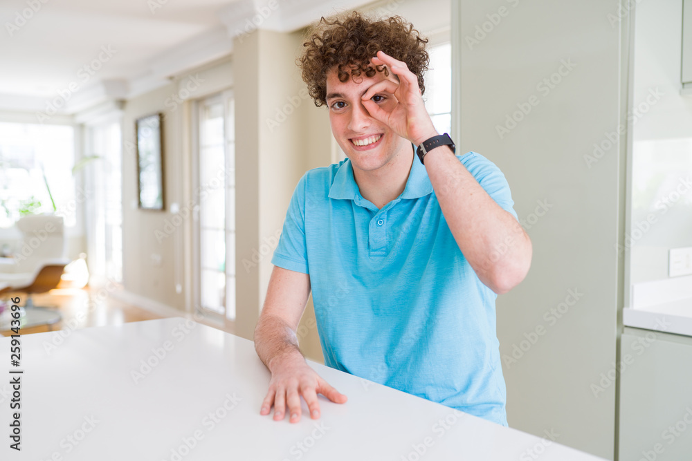Young handsome man wearing casual blue t-shirt at home doing ok gesture with hand smiling, eye looking through fingers with happy face. Young handsome man wearing casual blue t-shirt at home doing ok gesture with hand smiling, eye looking through fingers with happy face.