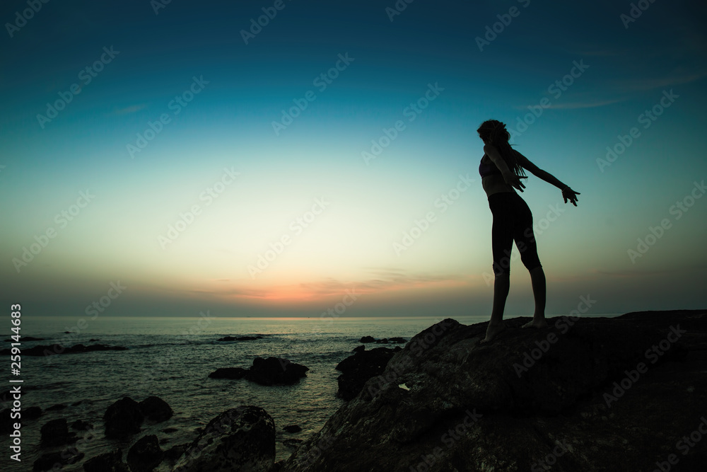 Silhouette of flexible yoga woman stands on the shore of the ocean at amazing evening.