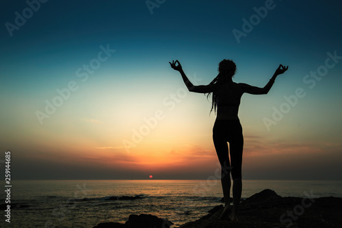 Silhouette of a flexible yoga woman stands on the shore of the evening ocean.