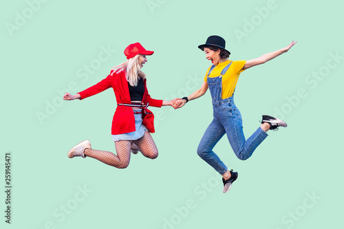 Full legs body size portrait of two happy screaming stylish hipster girls in fashionable clothes are jumping up in the air and celebrated a win. Indoor studio shot, isolated on green background