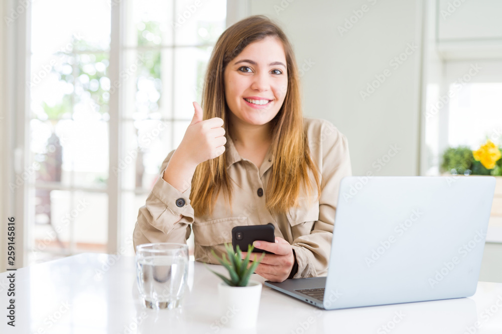 Beautiful young woman using smartphone and computer happy with big smile doing ok sign, thumb up with fingers, excellent sign Beautiful young woman using smartphone and computer happy with big smile doing ok sign, thumb up with fingers, excellent sign