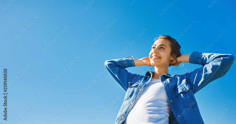 bottom view of a young smiling attractive woman in jeans clothes at sunny day on the blue sky background. joyful woman posing outdoors bottom view of a young smiling attractive woman in jeans clothes at sunny day on the blue sky background. joyful woman posing outdoors